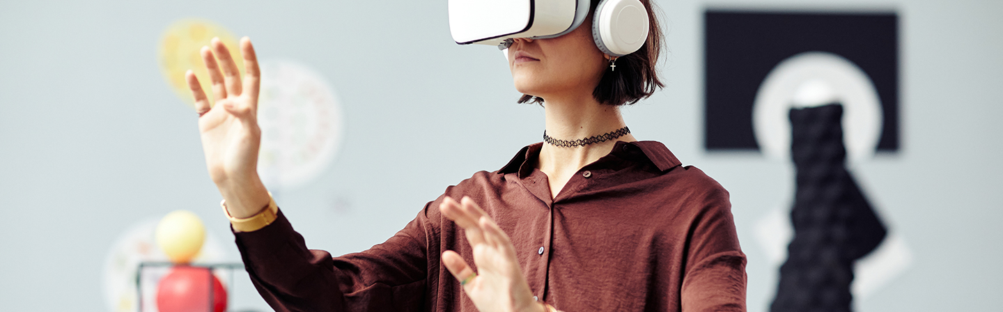 A young woman with dark brown medium-length hair is wearing white VR glasses and white headphones. She has both hands stretched forward groping.